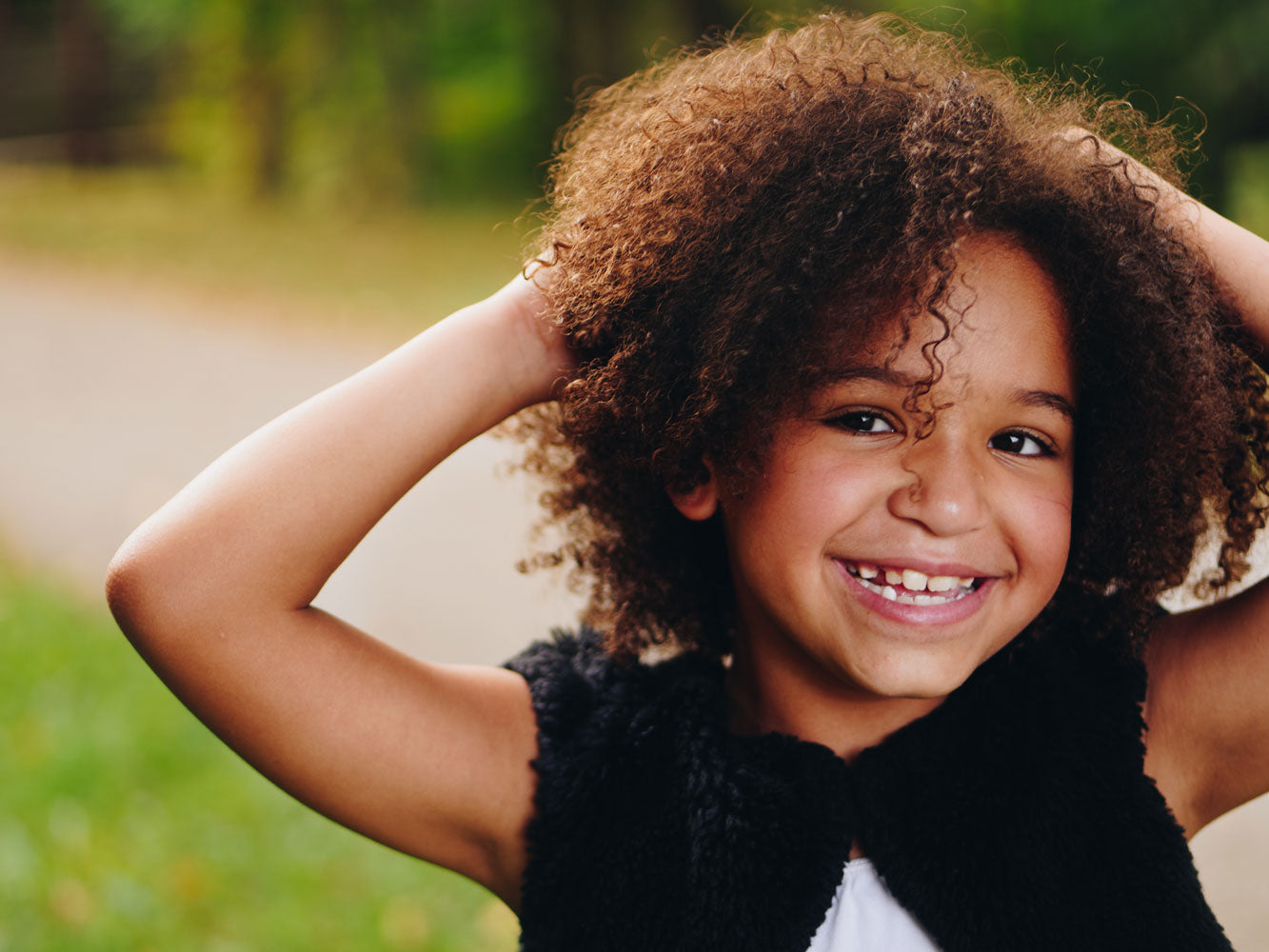 Face of smiling girl with curly hair standing outside with hands behind her head