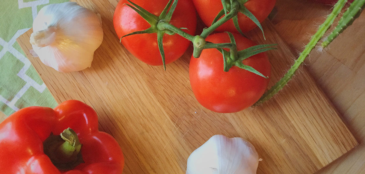Photo of a red pepper, two whole garlic heads and three tomatoes on a vine sitting on a wooden cutting board