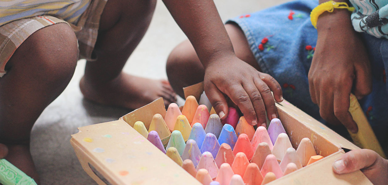 The hands of two children reaching for various colors of chalk in a box