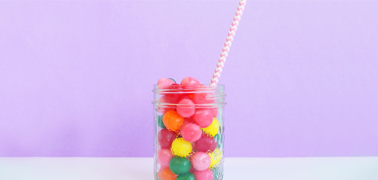 Glass jar filled with colorful candy and a white and pink straw against a purple background