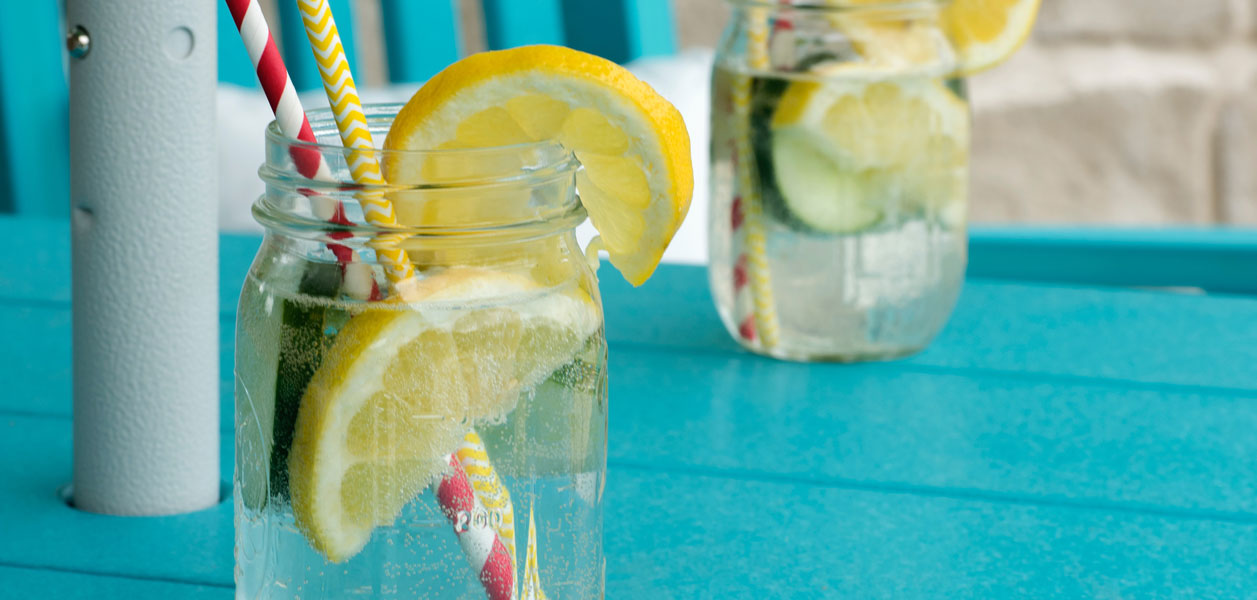 Two mason jars sitting on a blue table outdoors filled with sparkling water, lemons and cucumbers and two straws each